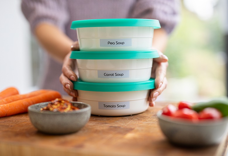 Ženska drži tri označene škatle Tupperware na deski za rezanje, na kateri so korenje, paradižnik in kumare