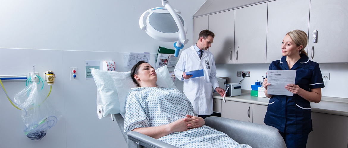 Pacientka leži na postelji, ob njej medicinska sestra z obrazcem, v ozadju zdravnik v beli uniformi