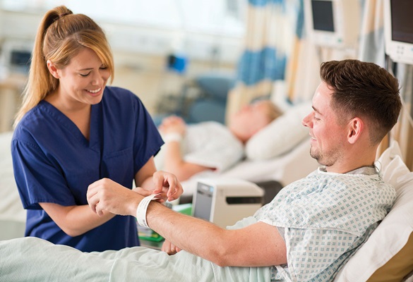 Medicinska sestra v modri uniformi pritrjuje pacientu, ki leži v postelji, ID-zapestnico